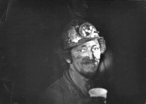Photograph of Miner Reg Pull underground wearing miner's helmet and holding a cup